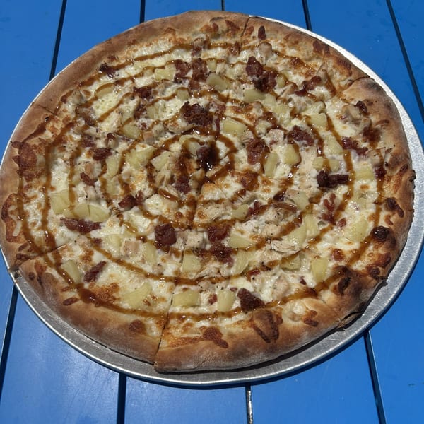 Shore Shack Pizza's BBQ chicken pizza with pineapple and a drizzle of BBQ sauce on a blue table.