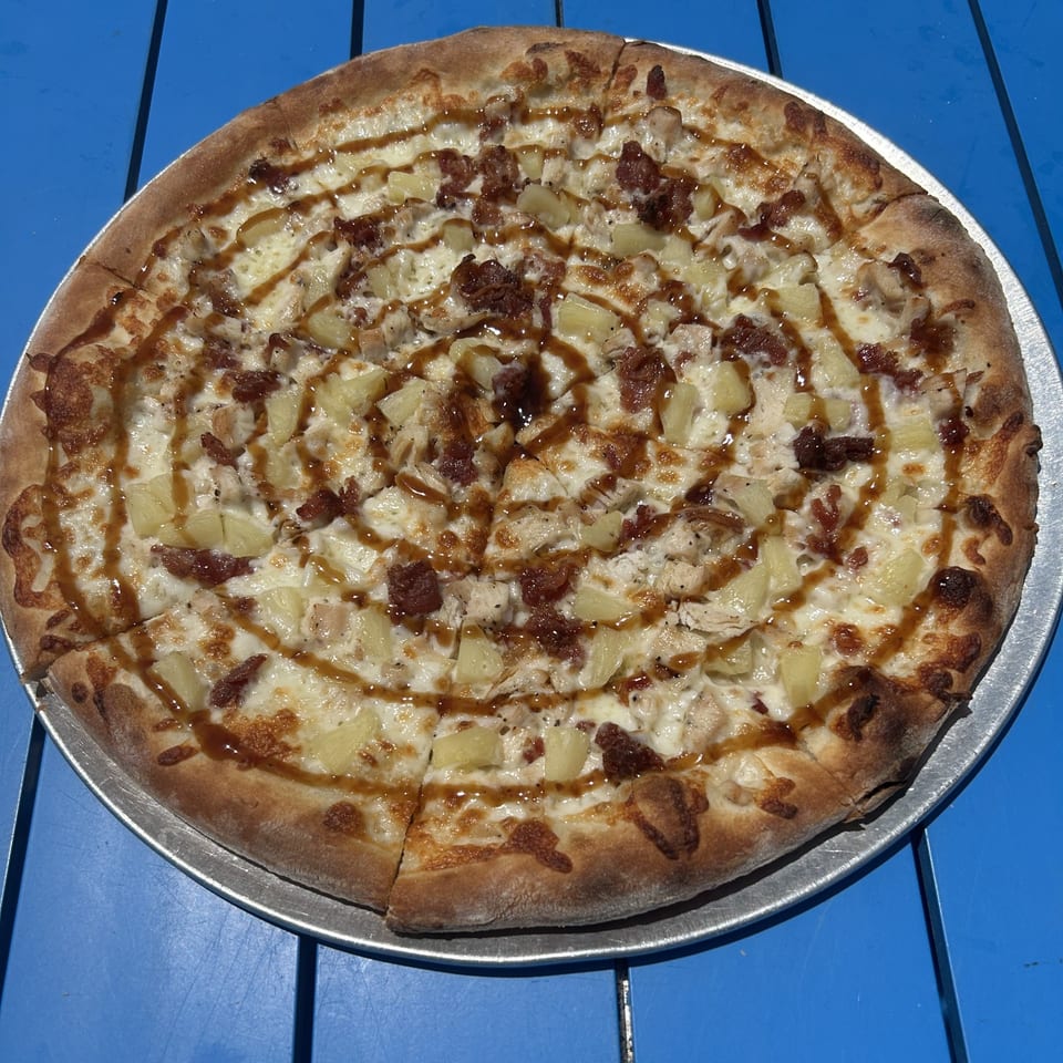 Shore Shack Pizza's BBQ chicken pizza with pineapple and a drizzle of BBQ sauce on a blue table.