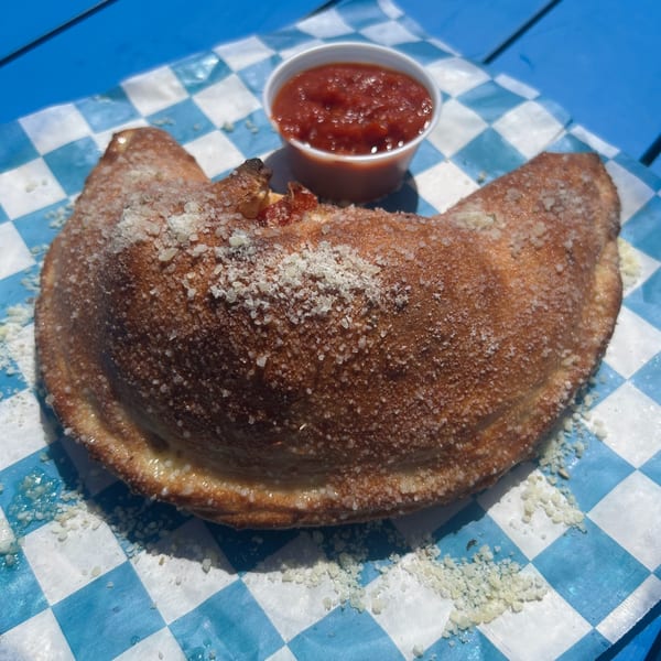 A golden-brown Shore Shack calzone, sprinkled with parmesan, rests on blue and white checkered paper with a side of marinara.