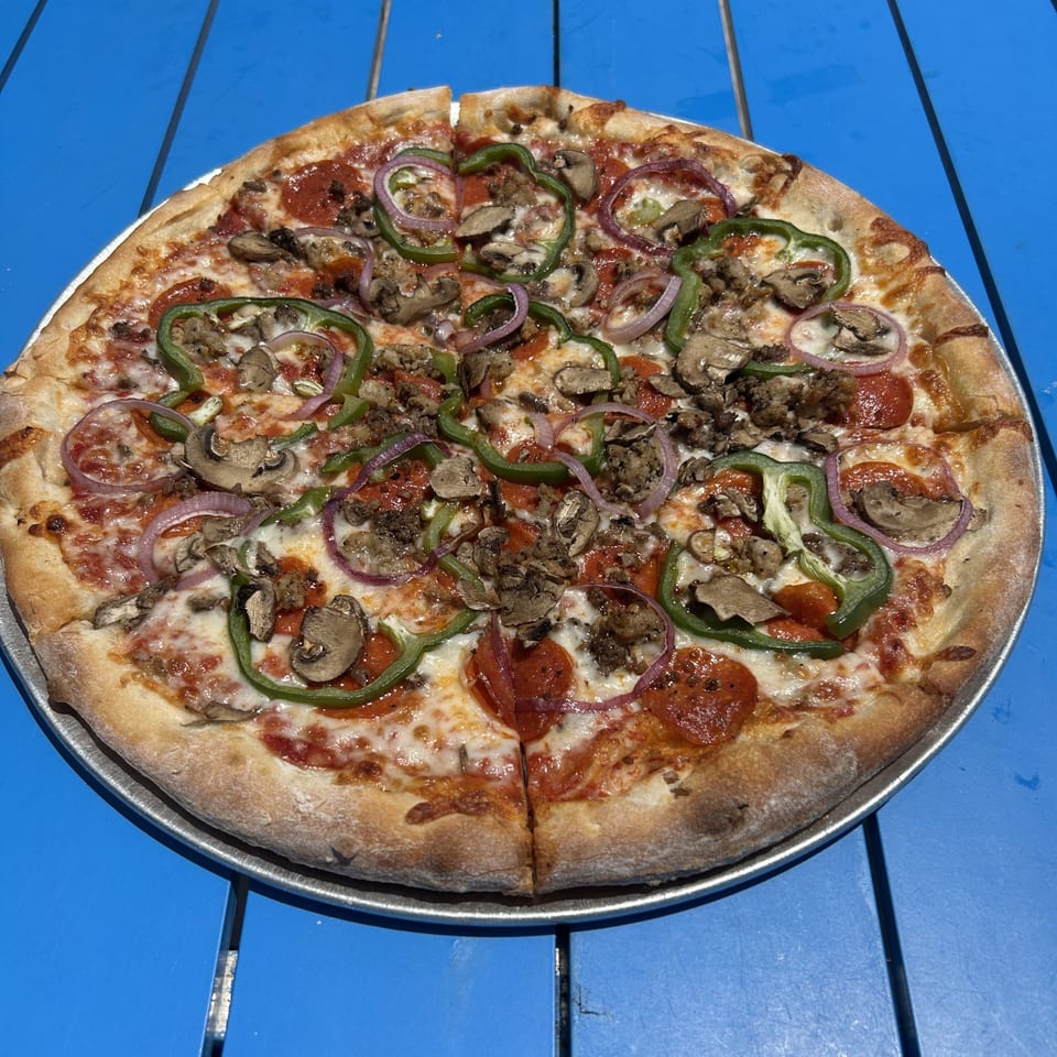 Shore Shack Pizza with pepperoni, sausage, mushrooms, green peppers, and red onions on a blue table.